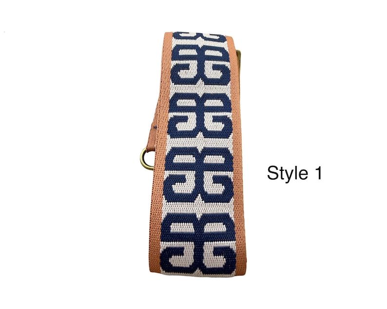 Pu&ograve; includere: Una tracolla tessuta con un motivo geometrico blu scuro ripetuto su sfondo grigio chiaro, bordata da bordi color marrone chiaro. La tracolla include componenti metallici e presenta la scritta Style 1 sul lato destro.