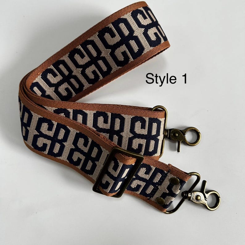 Pu&ograve; includere: Una tracolla per borsa in tessuto con un motivo geometrico blu navy ripetuto su sfondo beige e bordi color cuoio. Include un cursore in metallo regolabile e due moschettoni girevoli in ottone anticato. Il testo Style 1 &egrave; visibile.