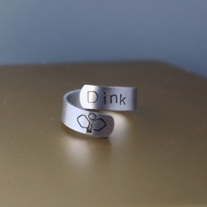 Puede incluir: Un anillo de plata con la palabra "Dink" grabada. El anillo también presenta una pequeña imagen de dos palas de pickleball y una pelota.