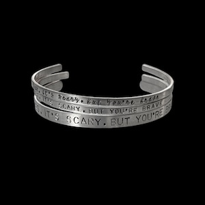 Pode incluir: Três pulseiras de punho prateadas empilhadas sobre um fundo preto. Cada pulseira tem o texto gravado: "It's scary, but you're brave."