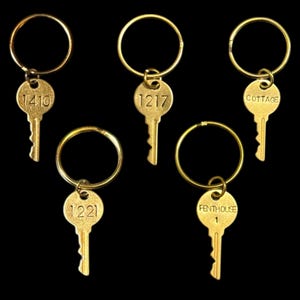 May include: Five vintage-style brass keys on keyrings. Each key has a different marking: "1410", "1217", "1221", "COTTAGE", and "PENTHOUSE 1". The keys are arranged against a black background.