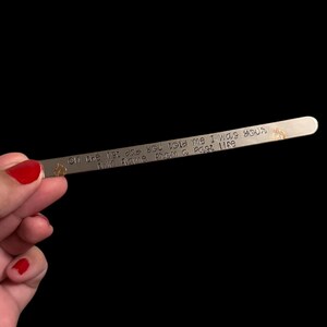 Puede incluir: Una barra de metal plateado con bordes redondeados, con texto estampado que dice: "On the 1st day you told me I was your twin flame from a Past life." Pequeños símbolos decorativos están estampados en la barra.