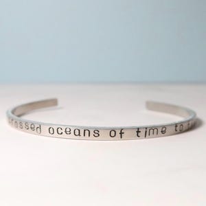 Può includere: Un bracciale a polsino color argento con la frase stampata "crossed oceans of time to". Il bracciale aperto è curvo e si appoggia su uno sfondo azzurro e bianco. Un gioiello semplice ed elegante.
