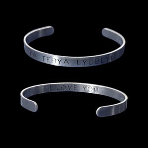 May include: Two silver-colored cuff bracelets. The top bracelet has the engraved text "YA TEBYA LYUBLYU". The bottom bracelet has the engraved text "I LOVE YOU". Both bracelets are set against a black background.