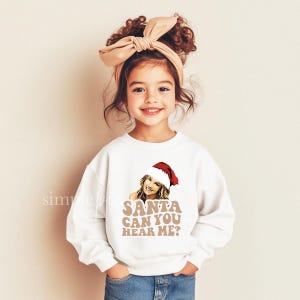 May include: A young girl in a white sweatshirt featuring a graphic of a woman in a Santa hat and the text "SANTA CAN YOU HEAR ME?". She is also wearing a beige headband and blue jeans.
