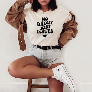 May include: A white t-shirt with black text that reads "No Daddy Just Issues" with a small heart below the text. The model is wearing the shirt with a brown corduroy jacket, denim shorts, and white sneakers.