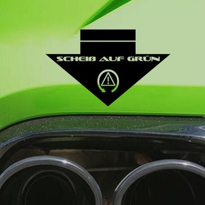 Puede incluir: Un coche verde con dos tubos de escape negros y una pegatina negra en el parachoques trasero que dice "SCHEIB AUF GRÜN" con un símbolo de un círculo con una flecha apuntando hacia la derecha.