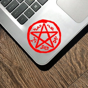 Pode incluir: Um pentagrama vermelho com um círculo ao redor, com símbolos escritos ao redor do círculo. O pentagrama está em um laptop prateado.