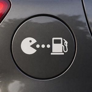 May include: A white Pac-Man character eating dots, followed by a white gas pump icon, on a dark gray car fuel door. The design is a playful take on refueling.