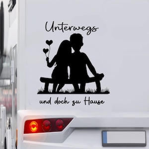 May include: Black silhouette decal of a couple sitting on a bench, with the text "Unterwegs und doch zu Hause". The woman holds heart-shaped balloons. The decal is applied to a white surface.