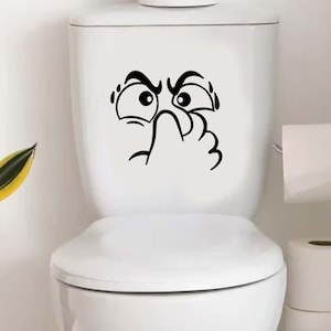 Könnte beinhalten: Eine weiße Toilette mit einem schwarzen Vinyl-Aufkleber eines Cartoon-Gesichts, das sich angewidert die Nase zuhält. Der Aufkleber ist auf dem Toilettentank zentriert. Toilettenpapierrollen sind rechts zu sehen, und links ist eine Pflanze.