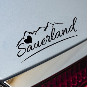 Könnte beinhalten: Schwarz-weißer Aufkleber mit einer Bergkette und einem Herzen, mit dem Text "Sauerland" in einer kursivschrift.