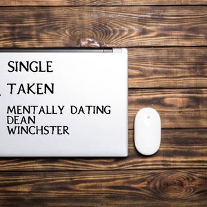 May include: A white laptop with a checklist on the screen. The checklist has three options: single, taken, and mentally dating Dean Winchester. The mentally dating Dean Winchester option is checked. A white computer mouse is on the wooden table next to the laptop.