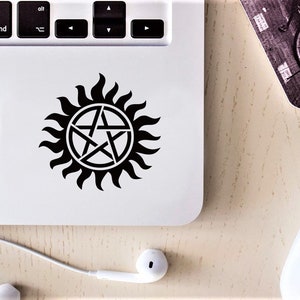 Op de afbeelding: Een zwart-witte sticker van een pentagram in een cirkel met vlammen eromheen. De sticker zit op een witte laptop.