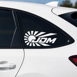 Può includere: Adesivo in vinile bianco con la scritta "JDM" in grassetto, con un design stilizzato del sole nascente. L'adesivo è applicato sul lunotto posteriore di un'auto bianca, presentando un'estetica pulita e moderna.