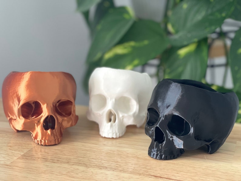 May include: Three 3D printed skull planters in different colors: copper, white, and black. The planters are made to look like human skulls and have a hollowed-out top for planting.
