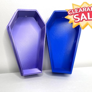 May include: Two coffin-shaped containers, one purple and one blue, are displayed. The blue container has a "CLEARANCE SALE" sticker. The containers are empty and have a simple, geometric design. They are likely decorative or storage items.