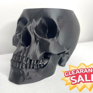 May include: A black skull-shaped planter with detailed facial features, including teeth and eye sockets. The planter is made of a solid material and has a hollow interior. A "CLEARANCE SALE" sticker is in the bottom right corner.