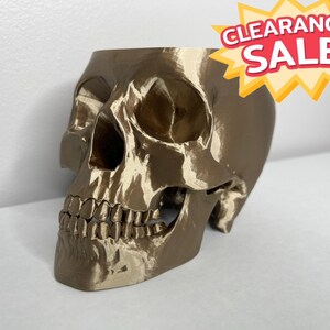 May include: A gold-coloured skull-shaped ornament. The skull features detailed teeth and eye sockets. A red and yellow "CLEARANCE SALE" sticker is in the upper right corner.