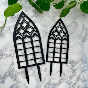 May include: Two black metal cake toppers shaped like gothic arched windows. The larger topper is approximately 10 inches tall and the smaller topper is approximately 6 inches tall.