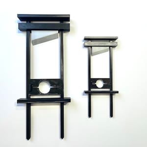 May include: Two black wooden guillotine models, one larger than the other, with a silver blade. The guillotines are on a white background.