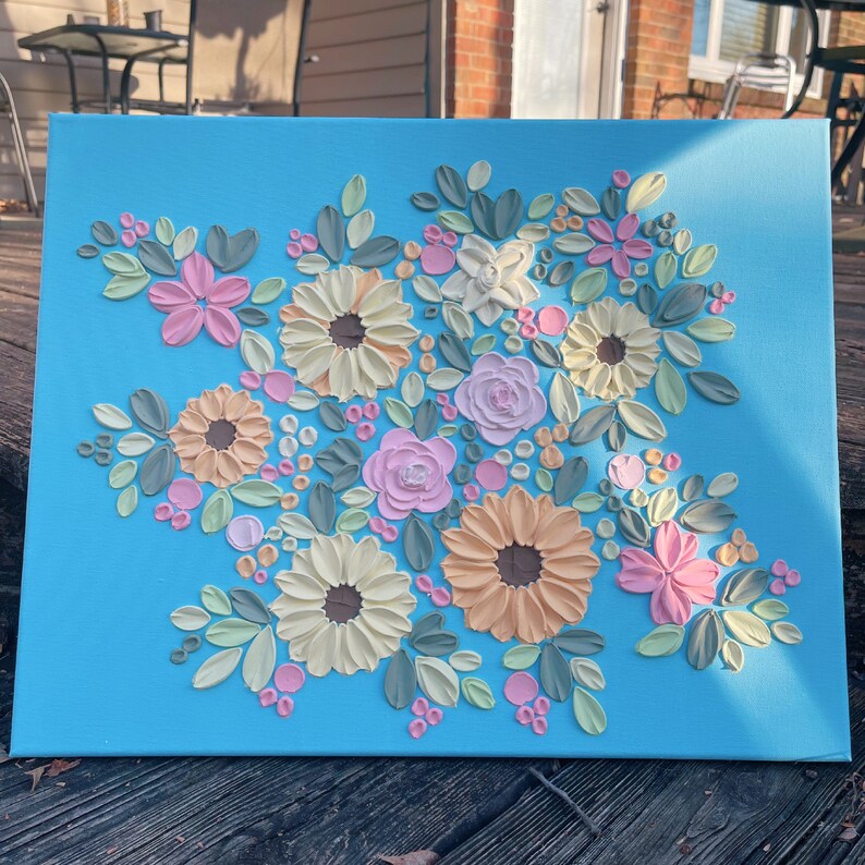 May include: A floral canvas art piece featuring a bouquet of textured flowers in shades of pink, peach, yellow, and white, with green leaves and pink berry accents, set against a bright blue background.