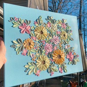 May include: A light blue canvas painting featuring a textured floral arrangement. The flowers are in shades of yellow, pink, and peach, with green and gray leaves. The artwork is held at an angle, showcasing the raised texture.