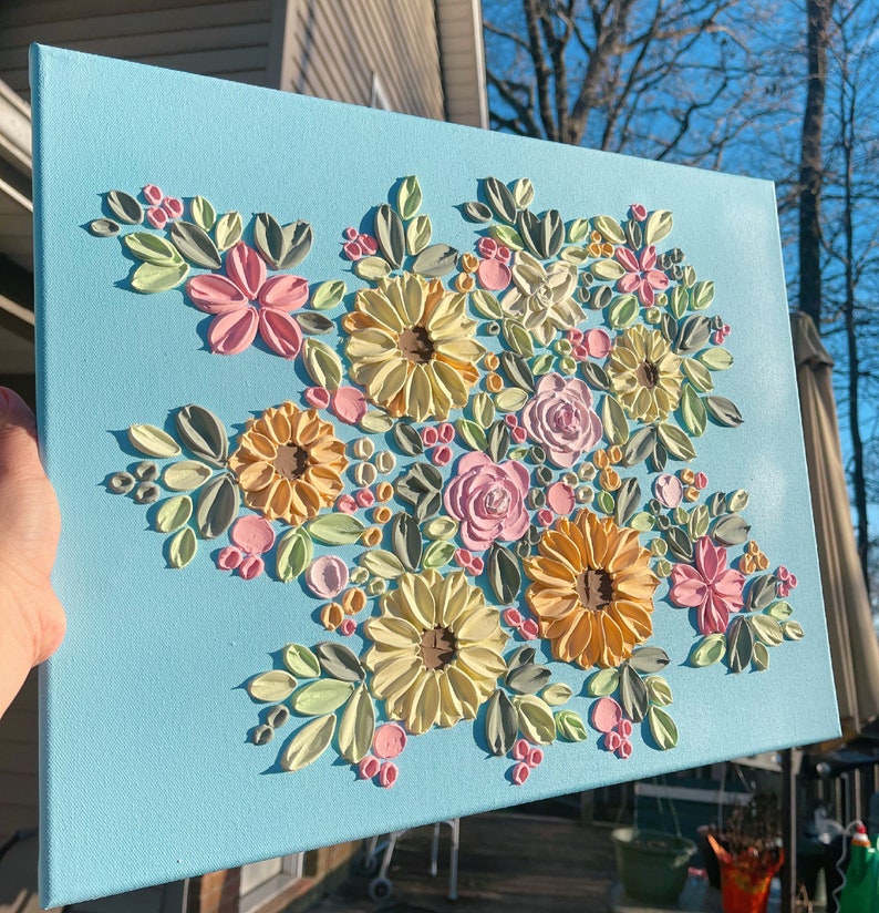 May include: A light blue canvas painting featuring a textured floral arrangement. The flowers are in shades of yellow, pink, and peach, with green and gray leaves. The artwork is held at an angle, showcasing the raised texture.