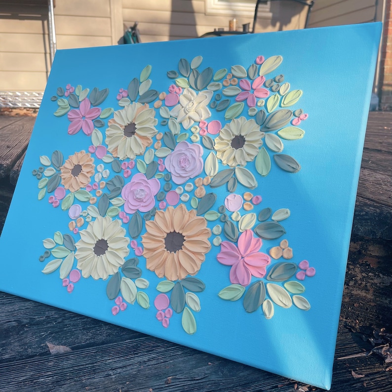May include: Square canvas art featuring a floral design with raised textures. The flowers are in shades of pink, yellow, and peach, with green leaves, set against a bright blue background. The artwork is displayed outdoors.
