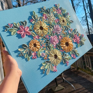 May include: A square canvas with a light blue background features a raised floral design. The flowers are in shades of yellow, pink, and peach, with green leaves. The artwork is displayed outdoors.