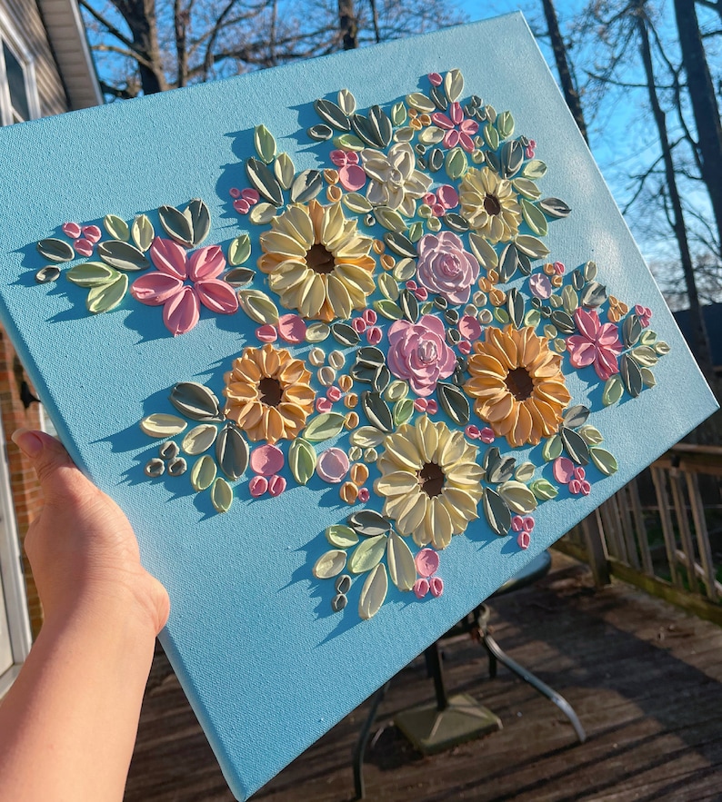 May include: A square canvas with a light blue background features a raised floral design. The flowers are in shades of yellow, pink, and peach, with green leaves. The artwork is displayed outdoors.