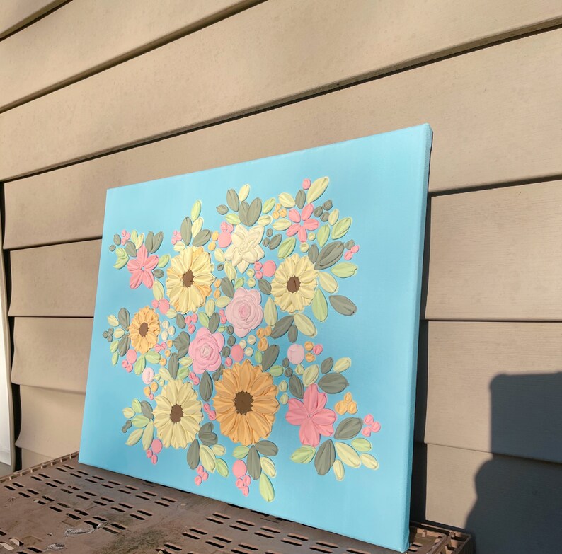 May include: A square canvas art piece featuring a floral design. The flowers are in shades of yellow, pink, and white, with green and gray leaves, set against a light blue background. The artwork has a textured, three-dimensional appearance.