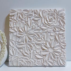 May include: White textured canvas wall art with a raised floral design. The flowers are in a variety of shapes and sizes, and they are all white.