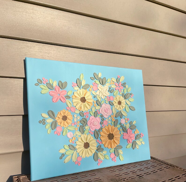 May include: A floral canvas art piece featuring a bouquet of textured flowers in shades of yellow, pink, and peach, with green and gray leaves, set against a light blue background. The artwork is displayed on a rectangular canvas.