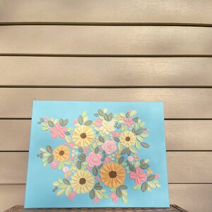 May include: A floral artwork on a light blue canvas. The painting features a bouquet of textured flowers in shades of yellow, pink, and peach, with green leaves. The flowers are arranged in a dense cluster, creating a vibrant and colorful composition.