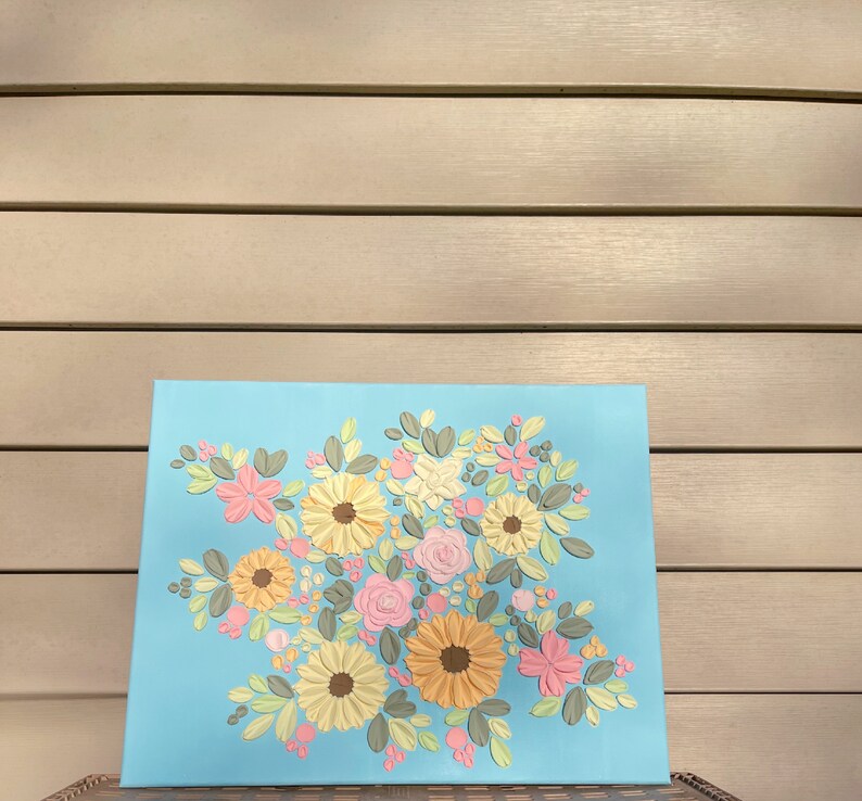 May include: A floral artwork on a light blue canvas. The painting features a bouquet of textured flowers in shades of yellow, pink, and peach, with green leaves. The flowers are arranged in a dense cluster, creating a vibrant and colorful composition.