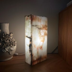 May include: A rectangular table lamp crafted from translucent stone with natural veining in shades of white, cream, and amber. The lamp emits a soft, warm glow from within, highlighting the organic patterns and textures of the stone material.