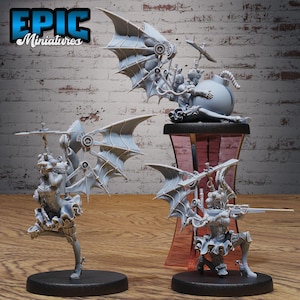 May include: Three grey miniature figurines with steampunk designs. One figure is seated on a bomb, another is kneeling with a rifle, and the third is standing. The figurines have wing-like structures and are on black bases. The text "EPIC Miniatures" is in the upper left corner.