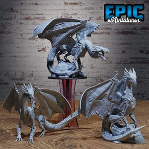 May include: Three grey dragon figurines with detailed wings and scales are displayed. One dragon is perched atop a stand, while the other two are positioned on a wooden surface. The logo "EPIC Miniatures" is visible in the upper right corner.