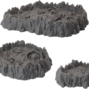 Puede incluir: Tres piezas de terreno grises y texturizadas para juegos de mesa. Las piezas tienen un aspecto rocoso y con cráteres, con tamaños y formas variables. La pieza más grande está en la parte superior, con dos piezas más pequeñas debajo. Diseñado para crear un paisaje realista.