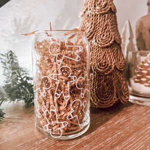 Puede incluir: Un tarro de vidrio transparente con un diseño de hombre de jengibre blanco. El tarro está lleno de papel de seda marrón y está sentado sobre una superficie de madera.