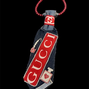 Peut inclure: Une cravate en jean avec un logo Gucci rouge et blanc et un collier de perles rouges. La cravate présente une étiquette Gucci rouge avec des lettres blanches, un charm de flacon de parfum et une épingle décorative avec une rose rouge. La cravate est ornée de strass.