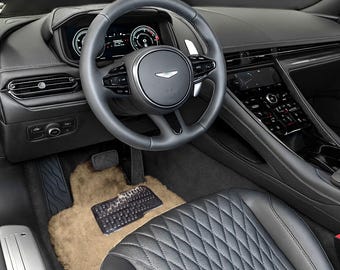 May include: Interior view of a luxury car featuring a black leather steering wheel, dashboard, and seats. The steering wheel has integrated controls and a silver emblem. A tan floor mat and a black rubber mat are visible in the footwell.