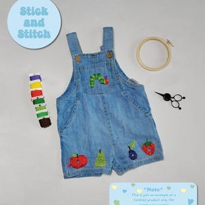 May include: Light blue denim overalls with embroidered fruits and a caterpillar. The overalls have a wooden embroidery hoop, scissors, and spools of thread. The text "Stick and Stitch" is in a light blue circle.