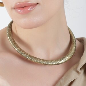 May include: A woman with curly brown hair wearing a gold choker necklace, gold hoop earrings, and a gold ring. She is wearing a light brown shirt.