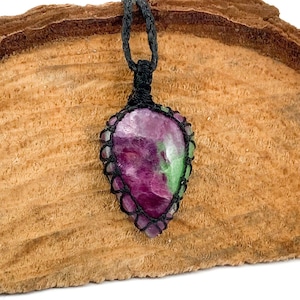Puede incluir: Un colgante de piedra preciosa verde y rosa en un collar de cordón negro. El colgante tiene forma de lágrima y está engastado en un nudo macramé.