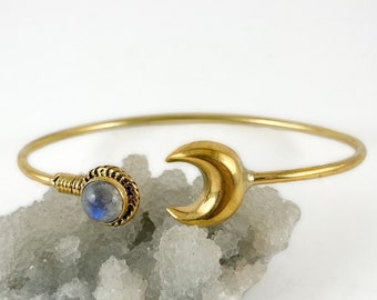May include: A gold bangle bracelet with a crescent moon charm and a moonstone cabochon. The bracelet is resting on a white crystal cluster.