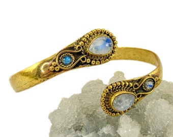 May include: A gold-toned cuff bracelet with two moonstone and one blue gemstone accents. The bracelet is on a white crystal surface.