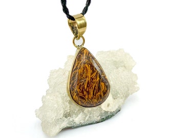 Op de afbeelding: Een gouden ketting met een hanger in de vorm van een traan, versierd met een bruine en gouden steen. De steen heeft een draaiende structuur.