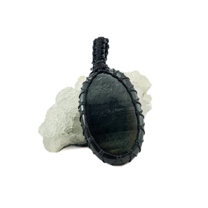 May include: Black oval gemstone pendant with a black macrame cord. The pendant is sitting on a white crystal cluster.